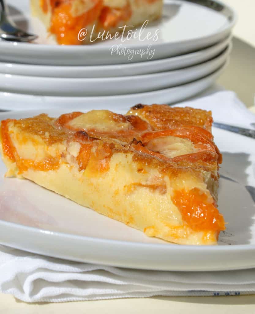 A close-up of a slice of apricot clafoutis on a white plate, placed on a folded napkin. The clafoutis has a custard-like texture with visible pieces of baked apricot. In the background, there are stacked white plates with more slices of the dessert. The scene is brightly lit, highlighting the golden, glossy surface of the dessert