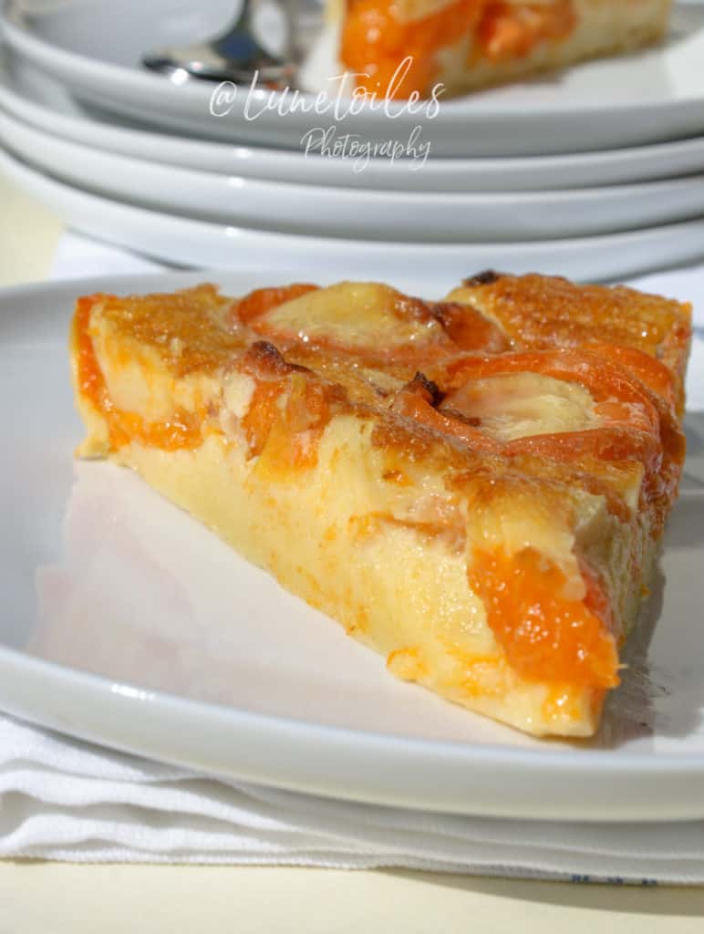 A close-up of a slice of apricot clafoutis served on a white plate, showcasing its golden-brown top and vibrant orange apricot halves embedded in a smooth, custard-like base. The plate rests on a white napkin, with another slice in soft focus stacked on a pile of plates in the background. The image is bright and sunlit, with the watermark "@Lunetoiles Photography" centered at the top