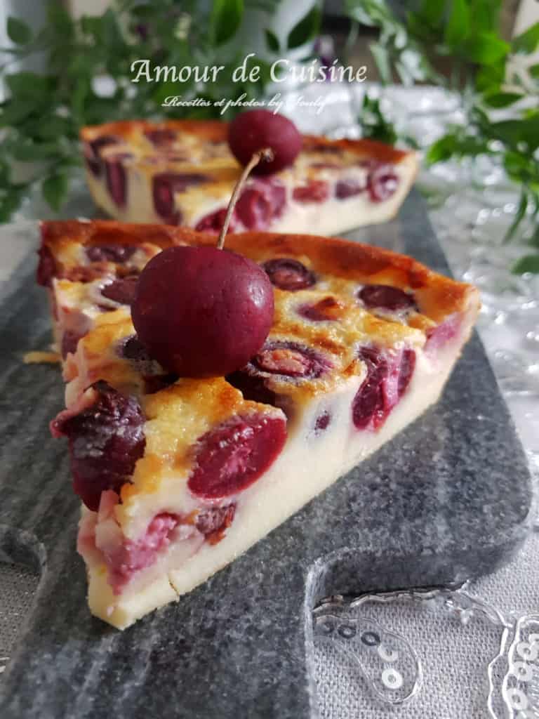 Slice of homemade cherry clafoutis with a golden top and juicy whole cherries, garnished with a fresh cherry on top, served on a stone serving board.