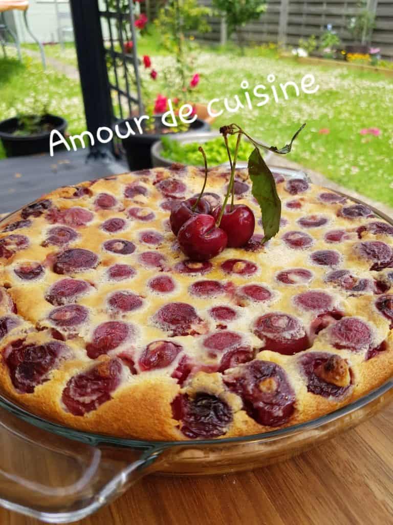 Whole homemade cherry clafoutis baked in a glass dish, golden and puffed with fresh cherries on top, garnished with a cherry stem, set on an outdoor table with a garden view