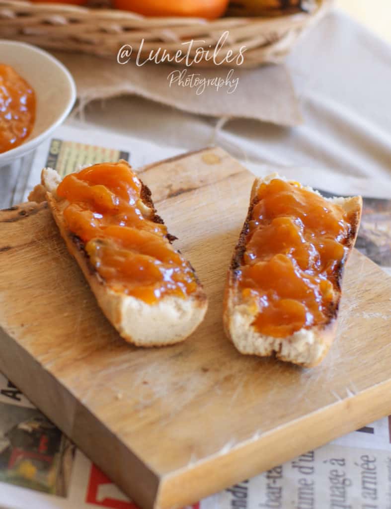 Two slices of baguette spread generously with glossy apricot jam, placed on a wooden board with a rustic background, including a bowl of jam and soft warm tones.