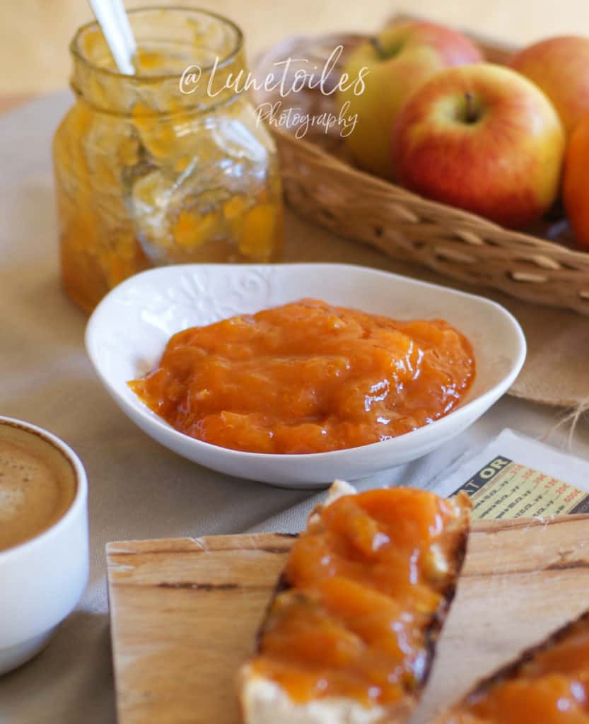 A bowl of thick, glossy apricot jam beside an open jar with a spoon, served next to slices of baguette topped with jam, a cup of coffee, and a basket of fresh apples in the background.