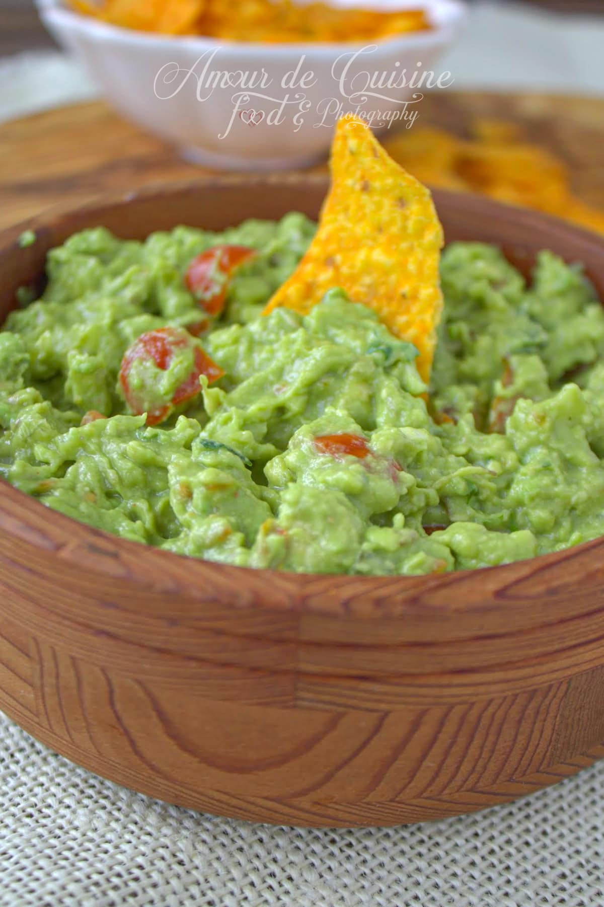 Guacamole maison crémeux dans un bol en bois, avec dés de tomate et tortilla plantée ; bol de nachos flou en arrière-plan — recette mexicaine authentique