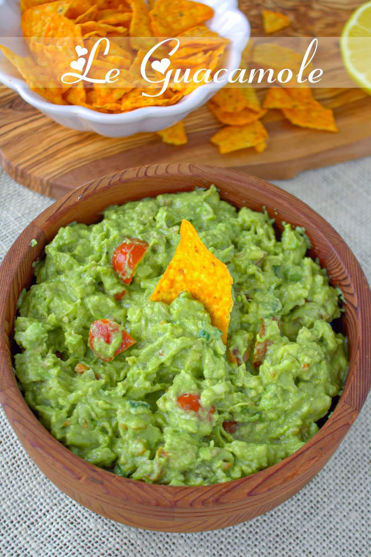 Bol en bois de guacamole maison aux dés de tomate, texture épaisse, tortilla plantée au centre ; nachos et quartier de citron en arrière-plan