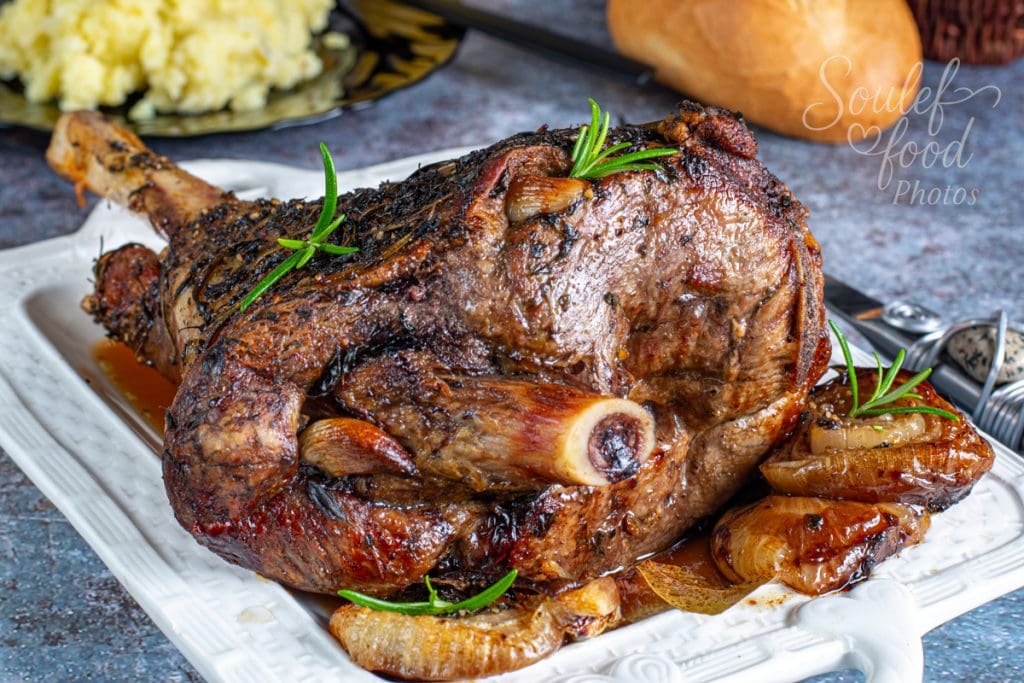 Gigot d'agneau rôti aux herbes servi avec des oignons caramélisés, présenté sur un plat blanc, accompagné d’une assiette de purée de pommes de terre en arrière-plan