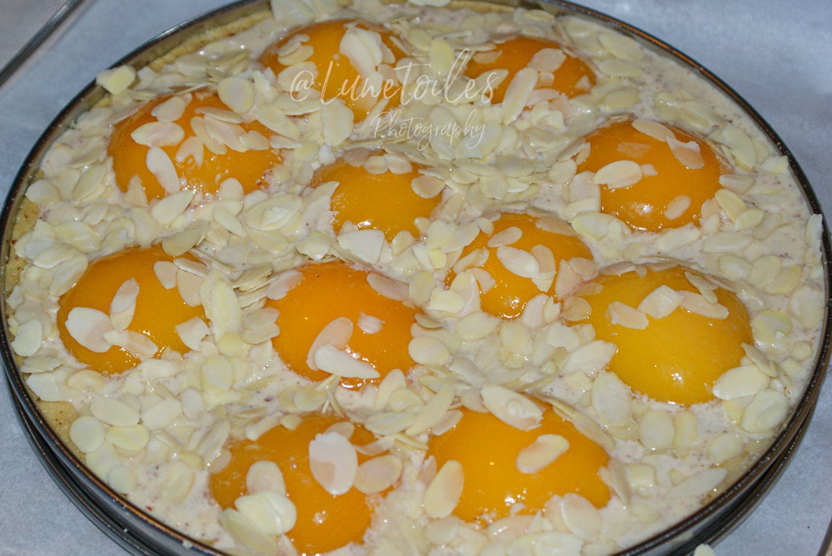 Tarte aux pêches amandine avant cuisson, garnie de demi-pêches au sirop, crème d’amande et amandes effilées.