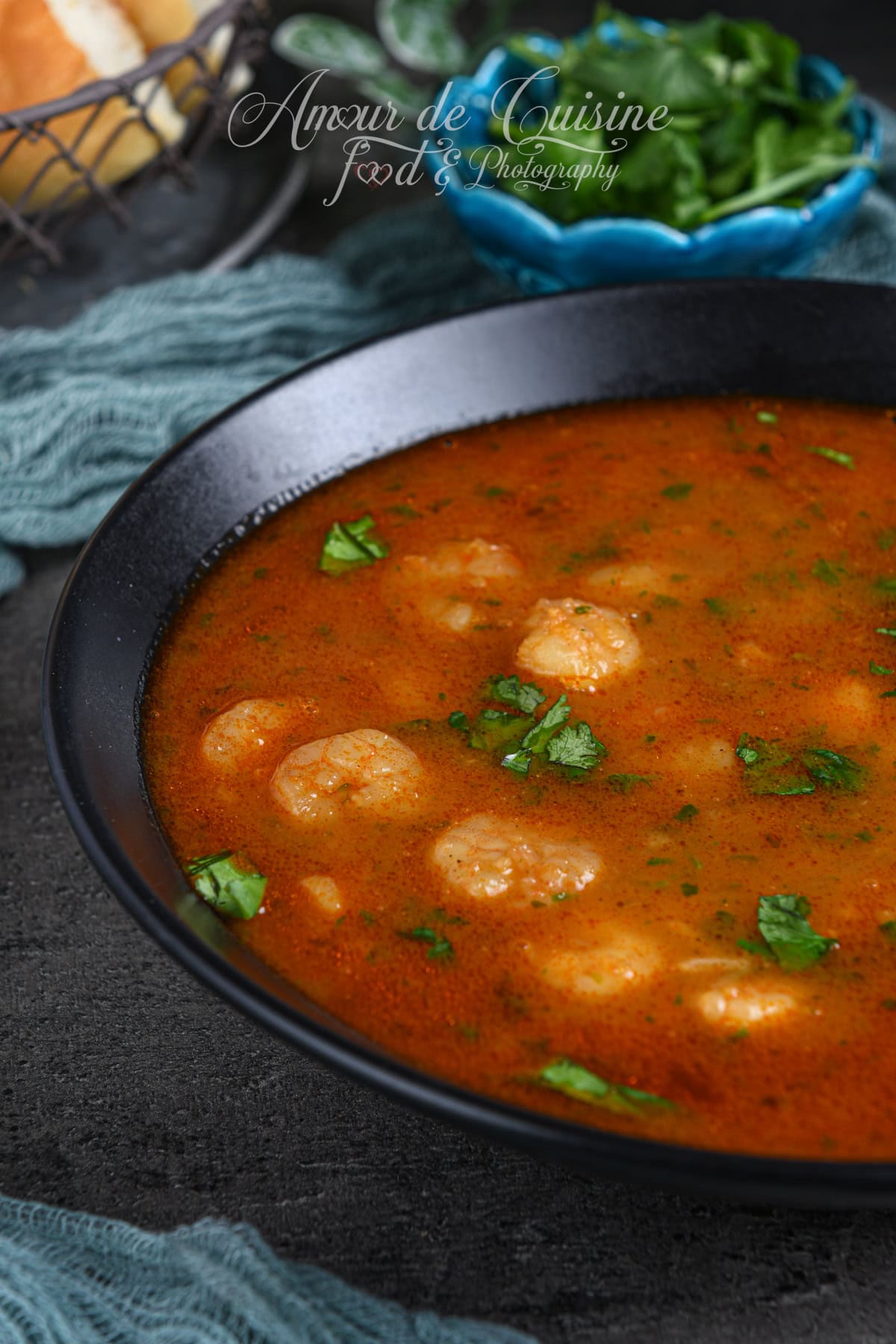 Gros plan d’une soupe de crevettes algérienne à la tomate, parsemée de coriandre, avec les crevettes bien visibles dans un bol noir sur surface sombre.