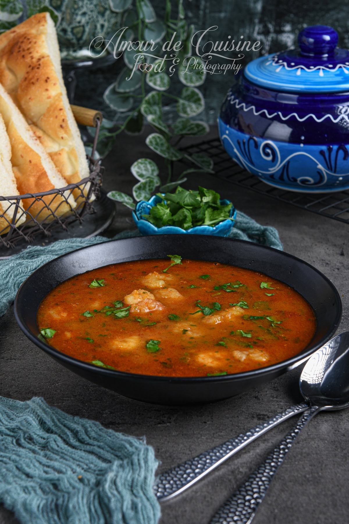 Bol noir de soupe de crevettes algérienne à la tomate, parsemée de coriandre, avec pain plat à côté et poterie bleue en arrière-plan.