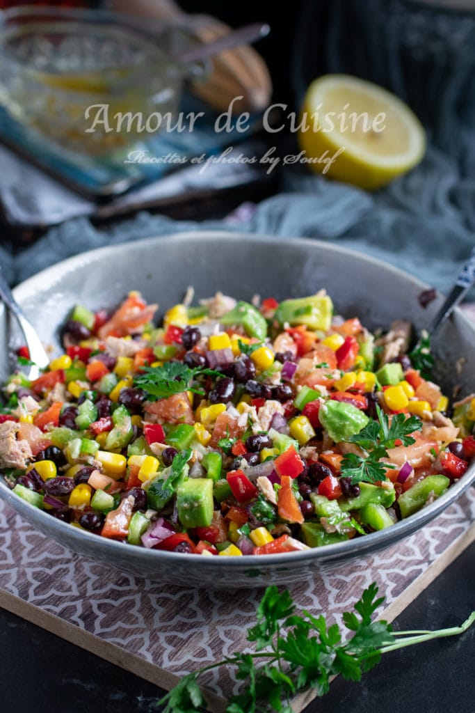 A vibrant, freshly mixed Mexican black bean and corn salad in a large bowl, filled with colorful diced vegetables including avocado, red onion, corn, tomatoes, bell peppers, and black beans. Garnished with parsley and set on a patterned board, with a halved lemon and dressing jar in the background