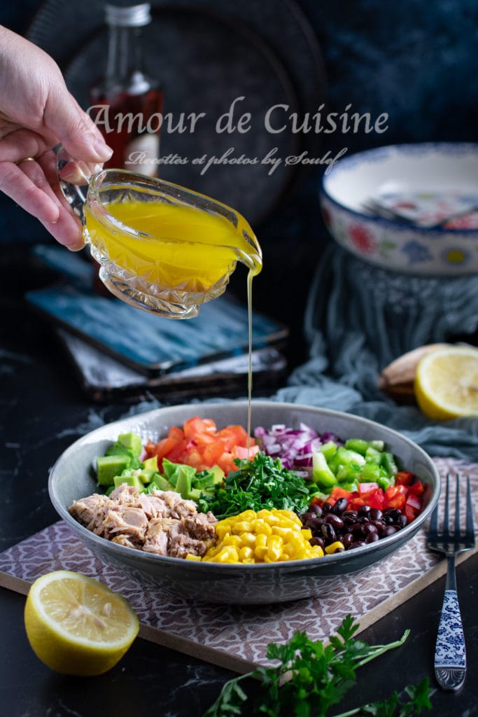 A hand is pouring a golden vinaigrette dressing from a glass jug onto a colorful Mexican salad in a bowl. The salad features neatly arranged ingredients including tuna, corn, black beans, avocado, tomatoes, red onion, green bell pepper, red bell pepper, and fresh parsley. A halved lemon and fresh herbs are placed nearby on a dark, elegant table setting