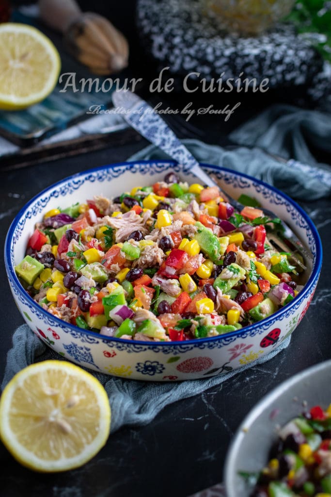 A vibrant bowl of Mexican salad featuring black beans, corn, diced avocado, red bell pepper, red onion, and tuna, garnished with fresh herbs. The salad is served in a decorative floral ceramic bowl with a blue rim, placed on a dark surface with a halved lemon beside it