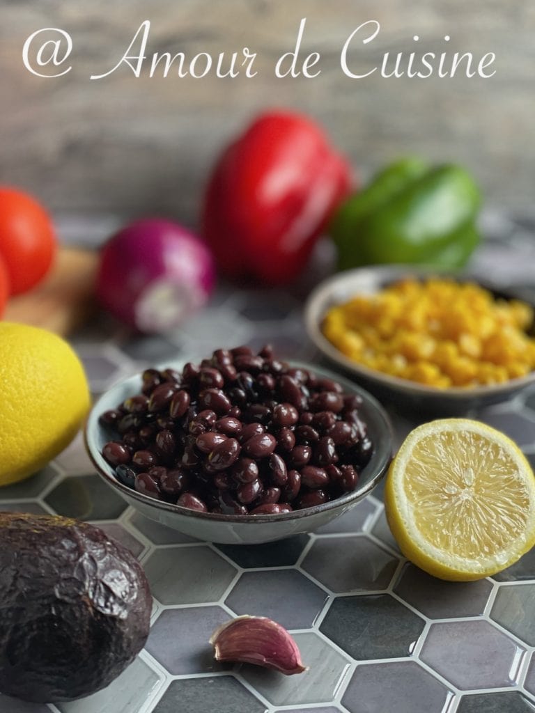 A close-up of fresh ingredients for a Mexican salad, including a bowl of black beans, a bowl of corn, a halved lemon, an avocado, a garlic clove, and various colorful vegetables like red onion, red and green bell peppers, and tomatoes in the background. The scene is set on a stylish hexagon tile surface