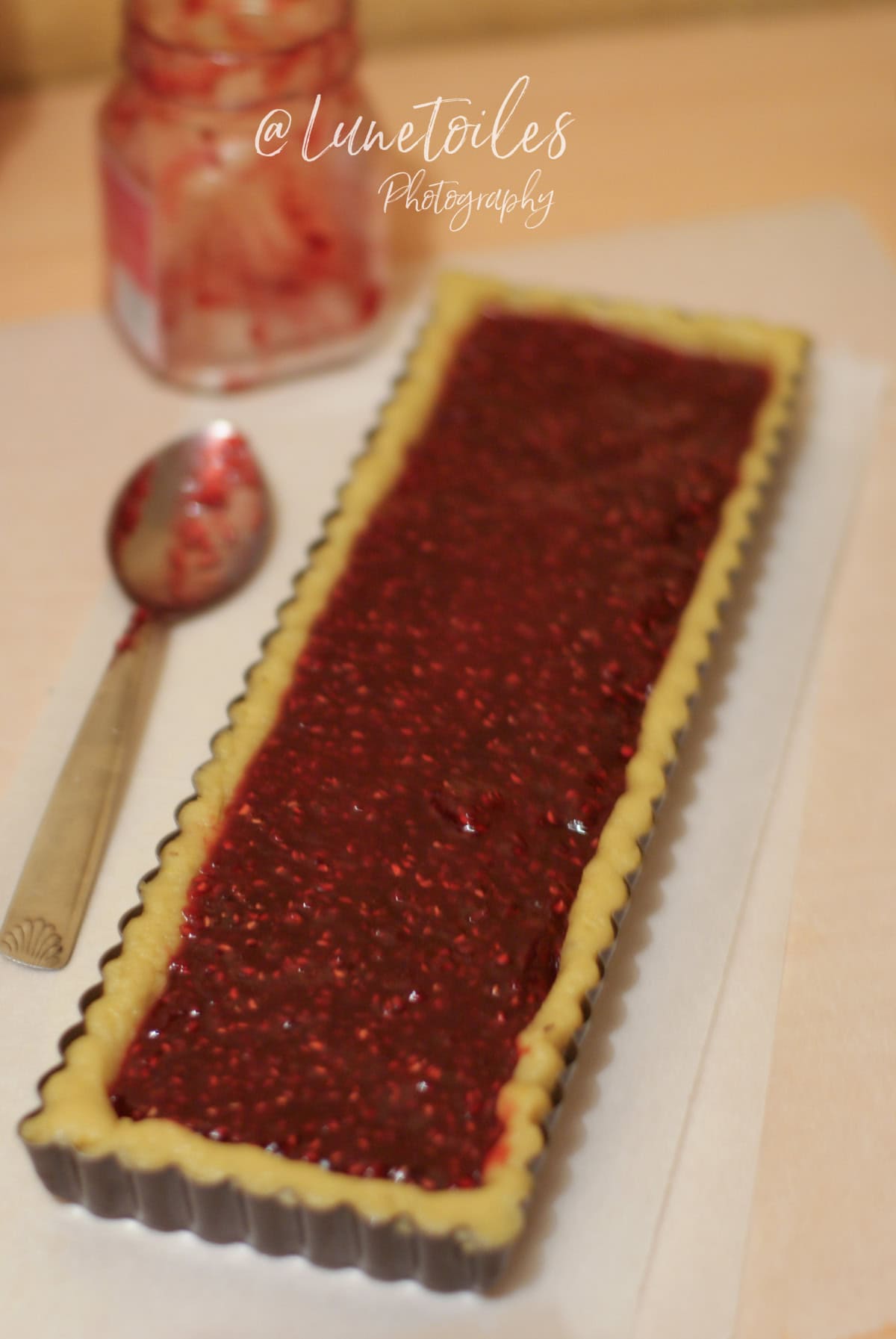 Fond de Linzer Torte dans un moule rectangulaire, garni généreusement de confiture de framboises, avec une cuillère et un pot de confiture en arrière-plan, photographie signée @Lunetoiles