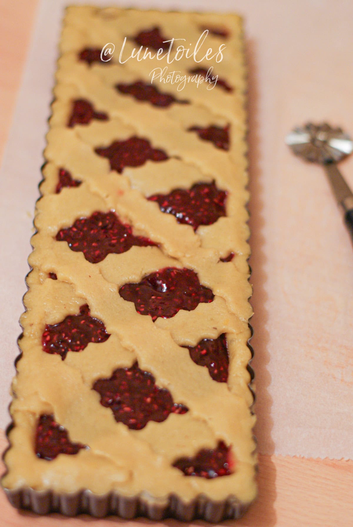 Linzer Torte crue dans un moule rectangulaire, garnie de confiture de framboises et décorée de croisillons de pâte sablée, avec une roulette à pâtisserie visible sur le côté, photographie signée @Lunetoiles