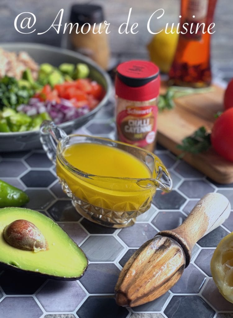 A vibrant kitchen scene showcasing ingredients for a zesty Mexican dressing, including a glass jug filled with golden vinaigrette, a halved ripe avocado, a wooden citrus juicer, a bottle of cayenne chili powder, and a salad bowl filled with chopped vegetables like tomatoes, red onion, avocado, and parsley in the background. Everything is arranged on a geometric tile surface for a fresh, rustic look