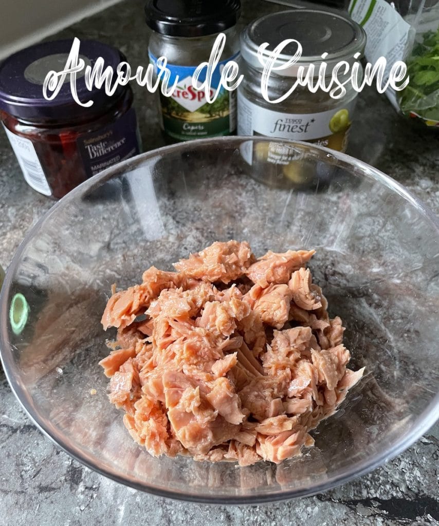 Bol en verre rempli de thon en boîte émietté sur un plan de travail, avec des bocaux de câpres et d’olives en arrière-plan.
