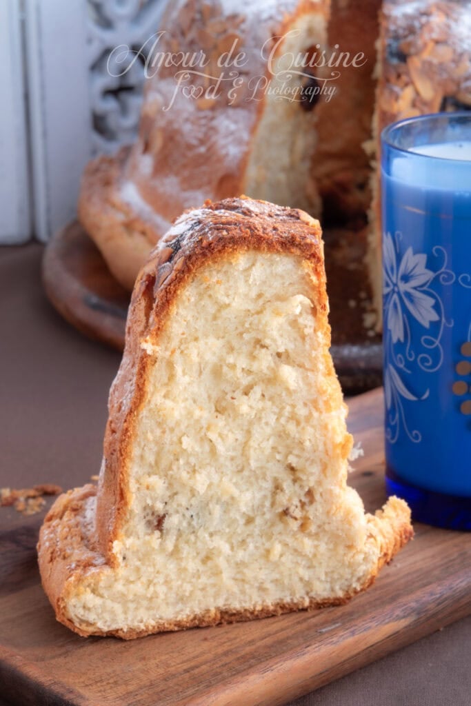 Grosse tranche de kouglof alsacien à la mie très moelleuse, posée sur une planche en bois, avec le kouglof en arrière-plan et un verre de lait bleu à côté.