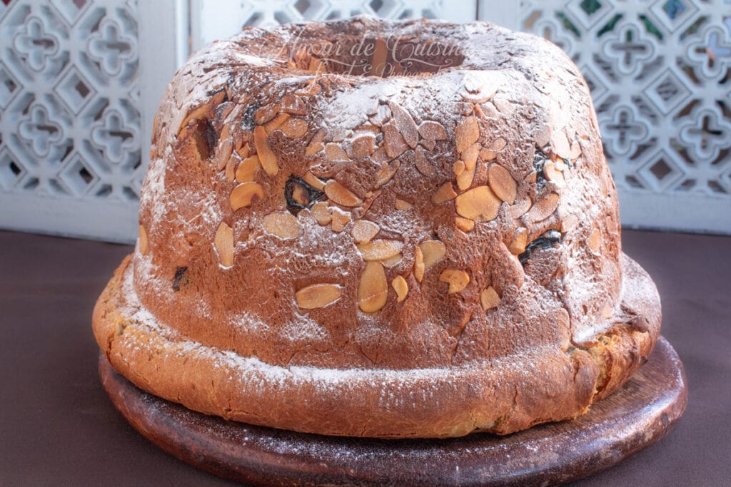 Kouglof alsacien entier, doré et bien gonflé, parsemé d’amandes effilées et légèrement saupoudré de sucre glace, posé sur une planche ronde en bois