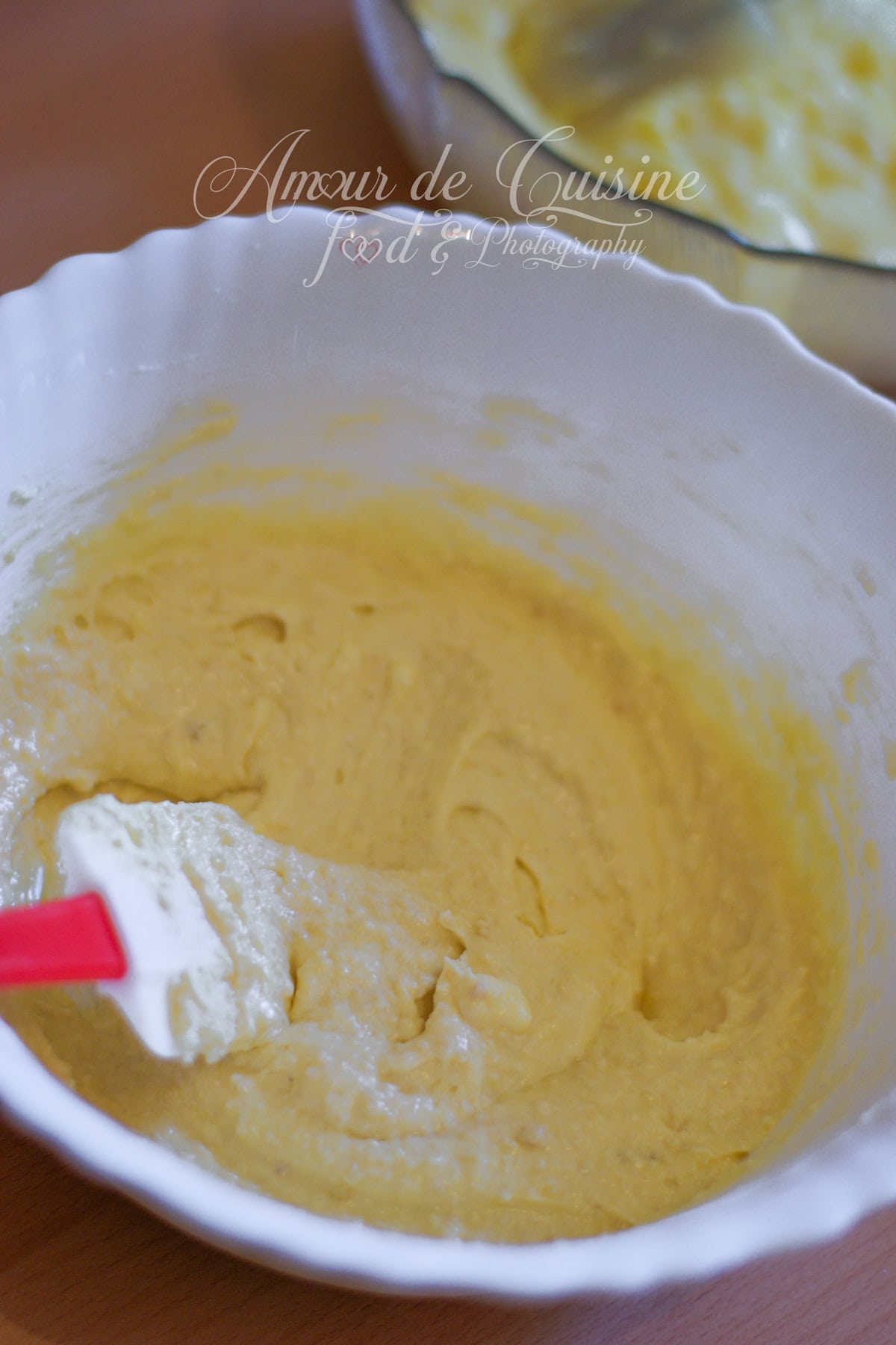 Crème d’amandes en cours de mélange dans un grand bol blanc, avec une spatule rouge, texture épaisse et lisse; un bol de crème pâtissière apparaît flou en arrière-plan.
