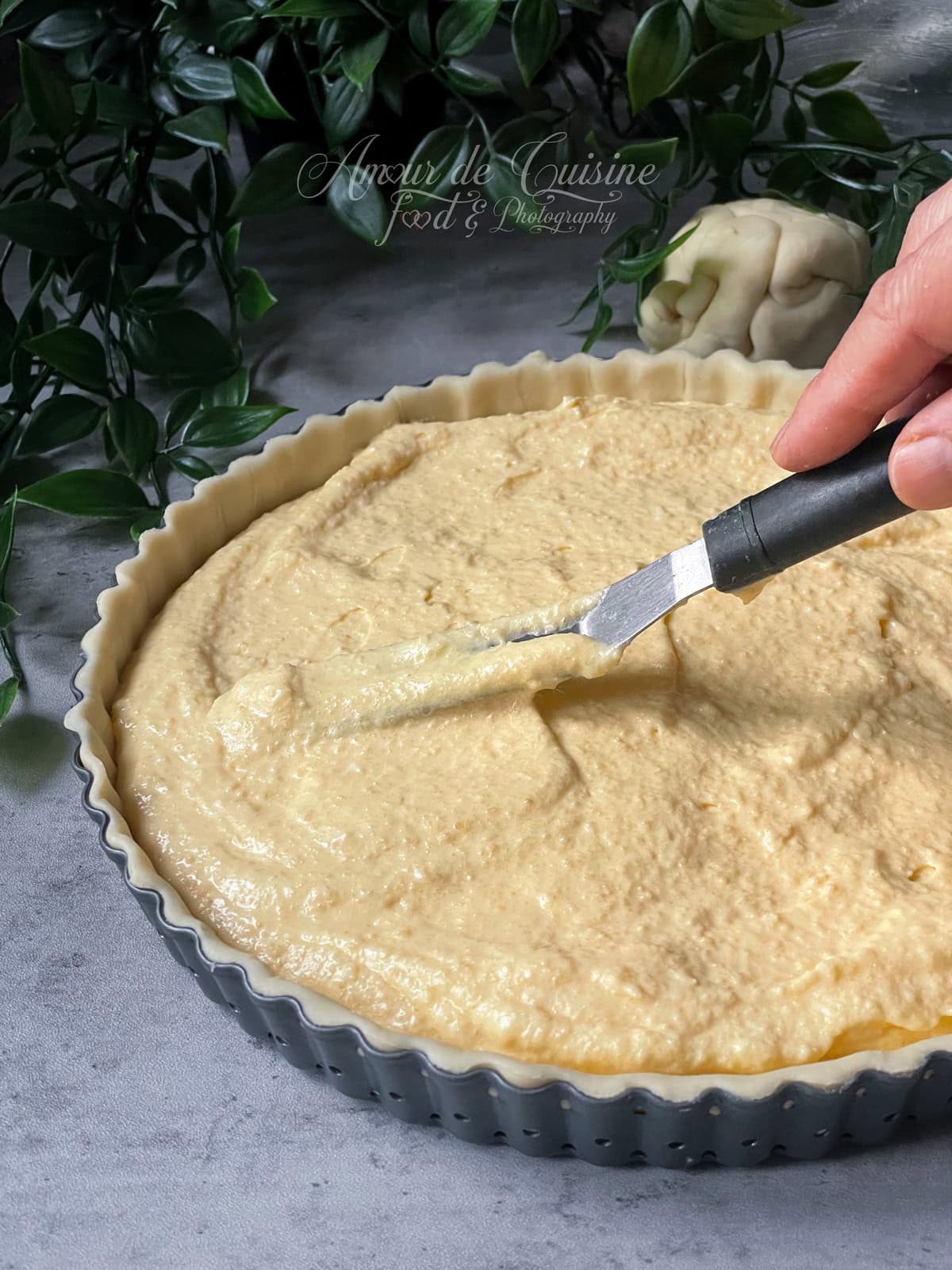 Fond de tarte dans un moule cannelé, garni de frangipane (melange crème d’amandes et creme patissiere) et lissé à la spatule avant cuisson, avec une main visible et des feuilles vertes en arrière-plan.
