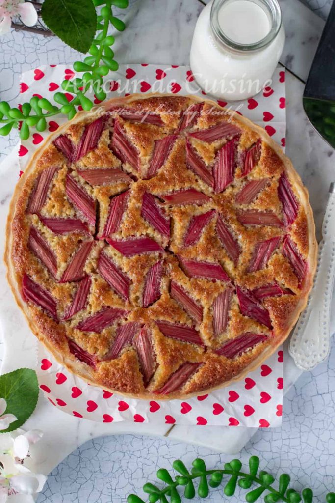 Overhead view of a beautifully baked rhubarb and almond cream tart with a golden crust and a striking geometric pattern of pink rhubarb on top. The tart is placed on a white board with red heart decorations, surrounded by greenery, flowers, and a bottle of milk, creating a fresh and inviting presentation