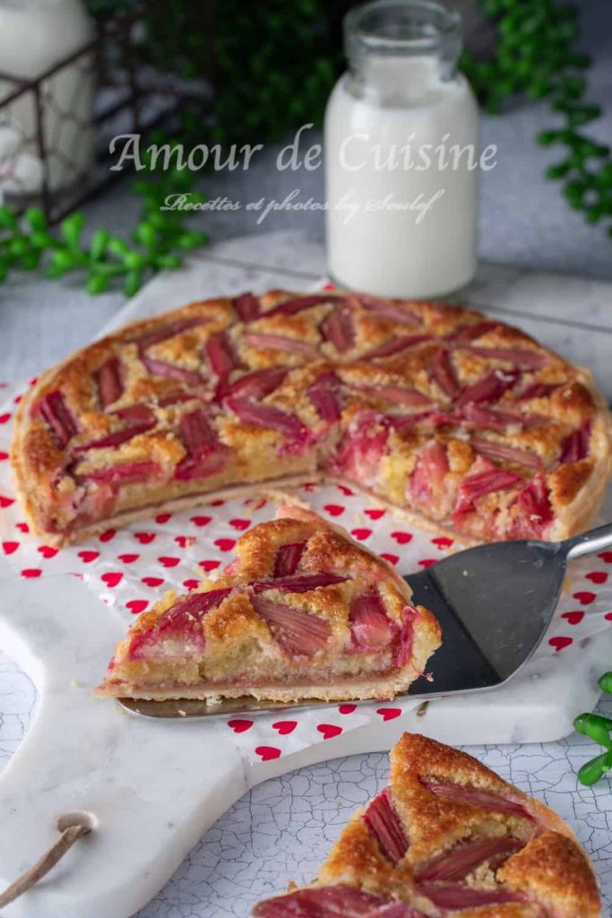 A freshly baked rhubarb frangipane tart on a white serving board with red heart patterns, featuring a slice being served with a spatula. The tart has a golden crust and decorative pieces of pink rhubarb on top, with a bottle of milk in the background