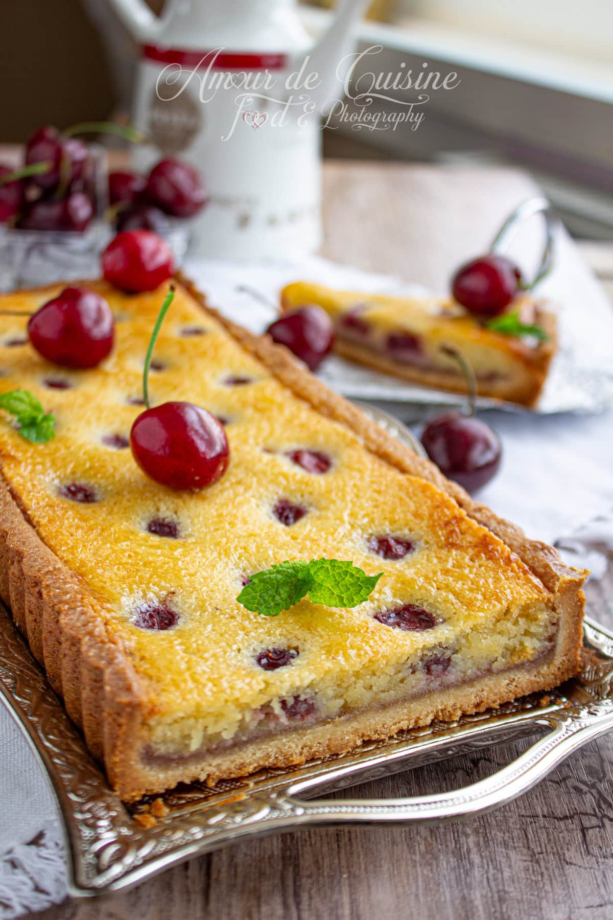 Tarte amandine aux cerises maison, rectangulaire, garnie de cerises entières et de crème d’amande dorée, présentée sur un plat en argent, avec feuilles de menthe fraîche en décoration