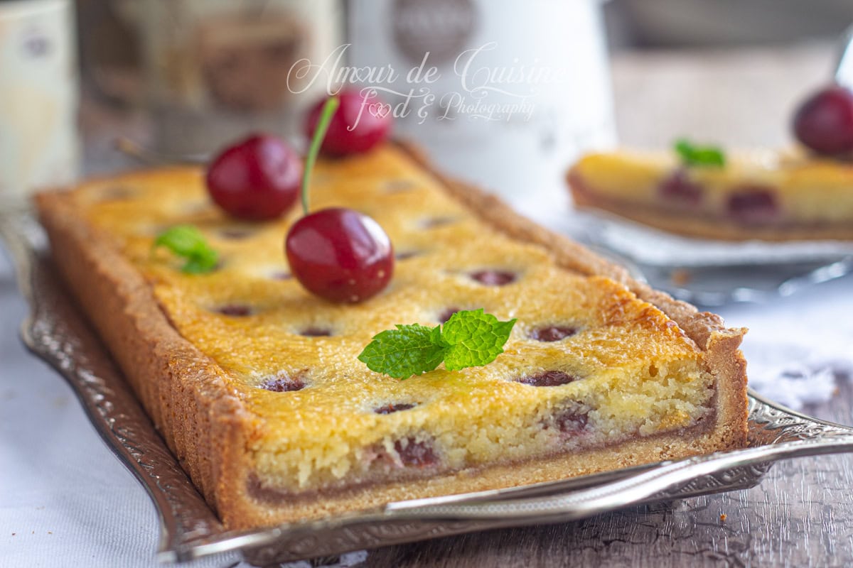Tarte amandine aux cerises rectangulaire, servie sur un plateau argenté, garnie de cerises entières et de feuilles de menthe, avec une pâte dorée et une crème d’amande moelleuse