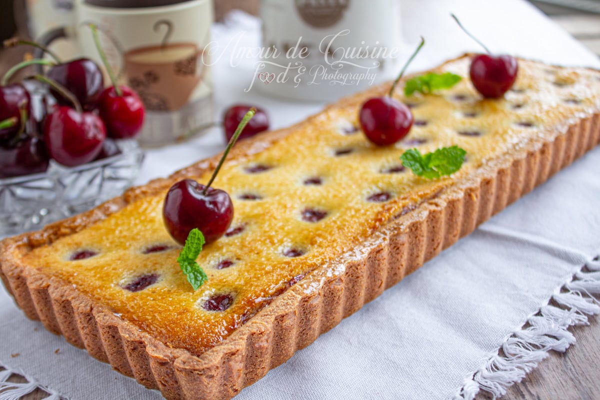Tarte amandine aux cerises rectangulaire, dorée au four, décorée de cerises fraîches et de feuilles de menthe, posée sur une nappe blanche avec des tasses en arrière-plan.