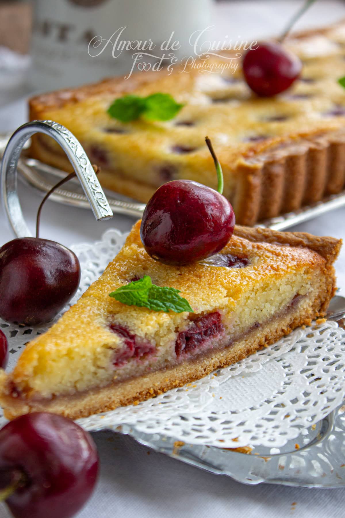 Part de tarte amandine aux cerises sur un plateau argenté, décorée d’une cerise fraîche et d’une feuille de menthe, avec pâte sablée dorée et crème d’amande moelleuse