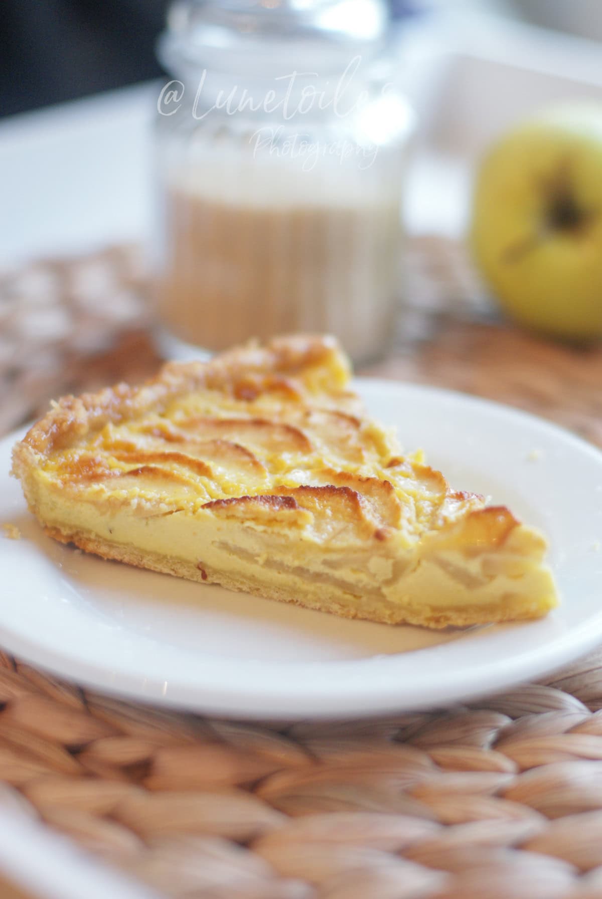 Slice of apple and mascarpone tart, golden and creamy, served on a white plate with a blurred drink glass and an apple in the background.
