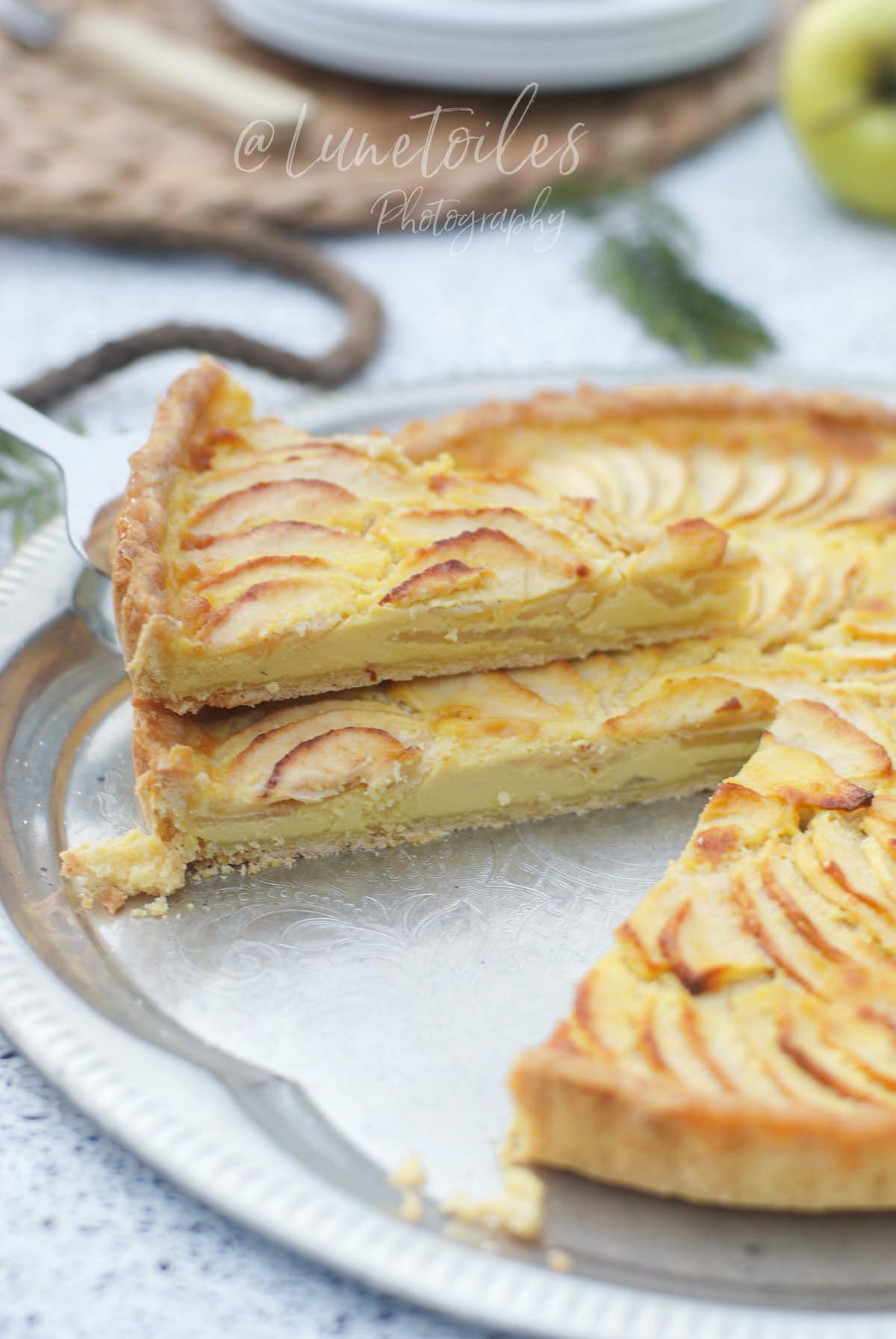 Golden apple and mascarpone tart sliced on a silver tray, with one slice lifted to reveal the creamy filling and thin apple layers.