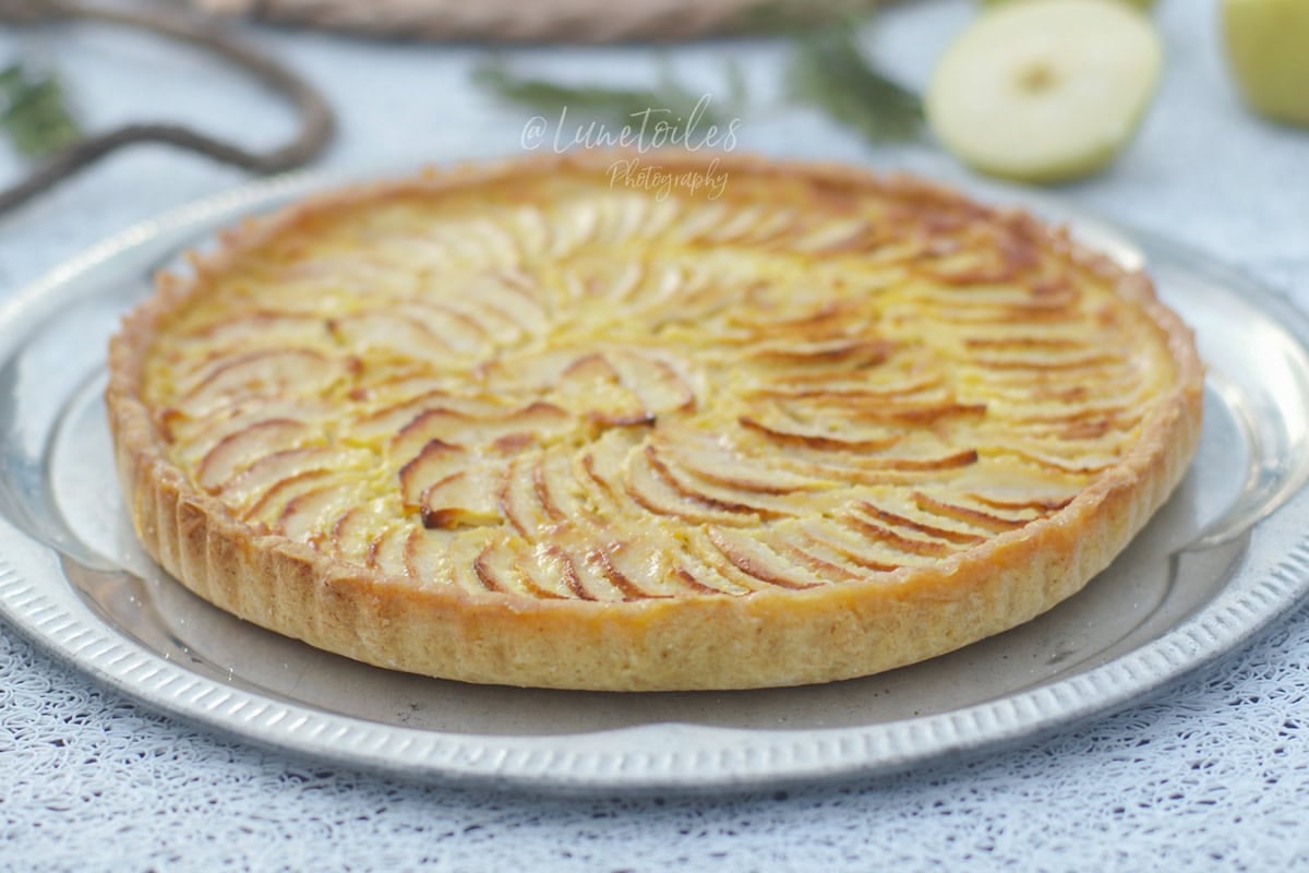 Whole apple and mascarpone tart, golden and crisp, topped with apple slices arranged in a rosette, presented on a silver tray with half an apple in the background.