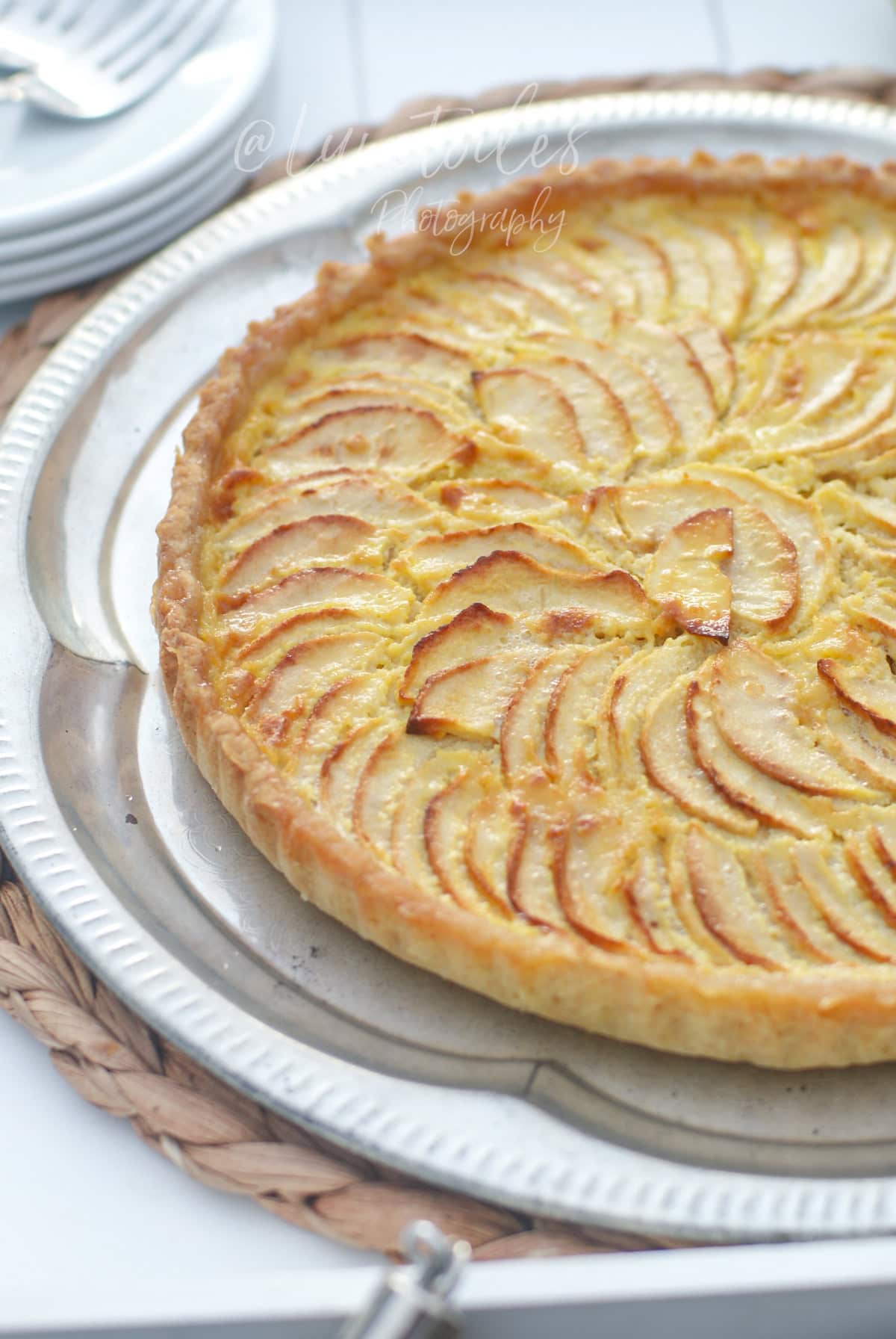 Whole apple and mascarpone tart, golden and glossy, topped with apple slices arranged in a rosette, set on a silver tray with plates in the background.