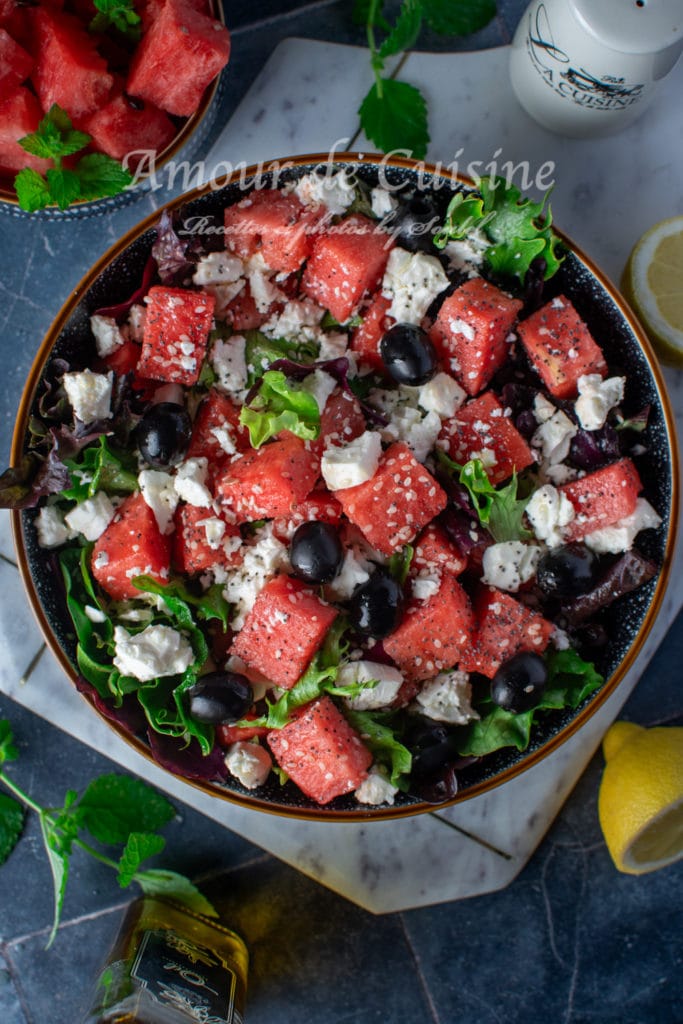 Top view of a watermelon feta salad with black olives, fresh greens, sesame seeds, and crumbled cheese in a black bowl, surrounded by lemon slices, mint, and olive oil on a dark surface