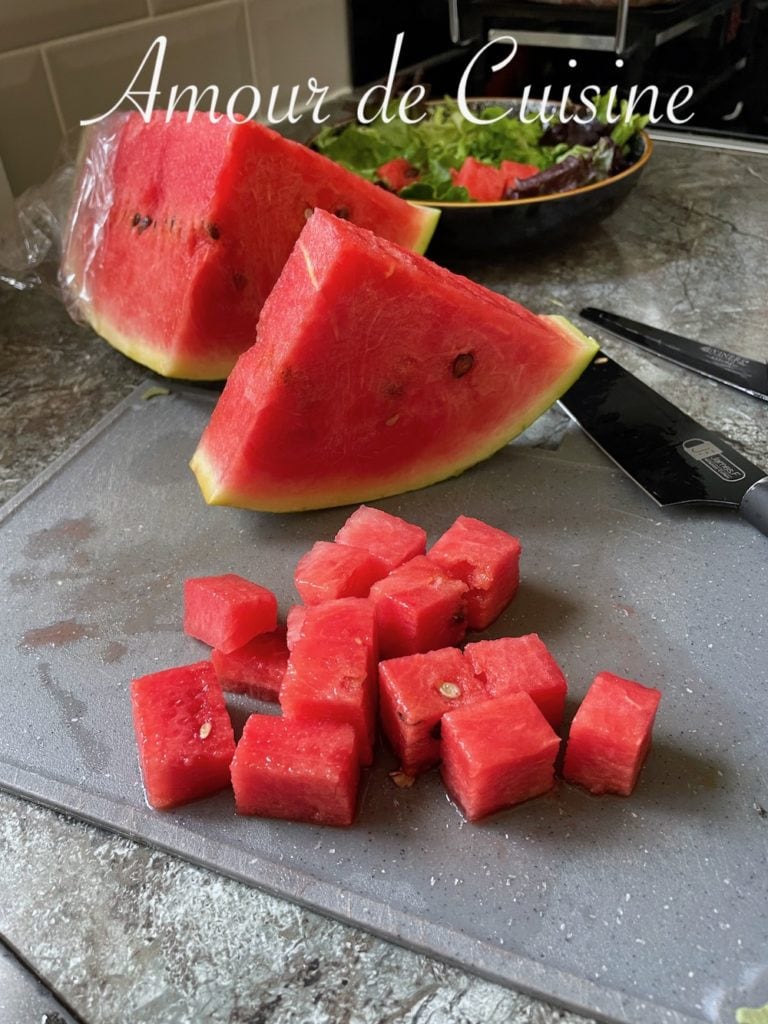 Cubed watermelon on a cutting board with large wedges in the background, next to a knife and a bowl of salad greens ready for assembling a watermelon feta salad