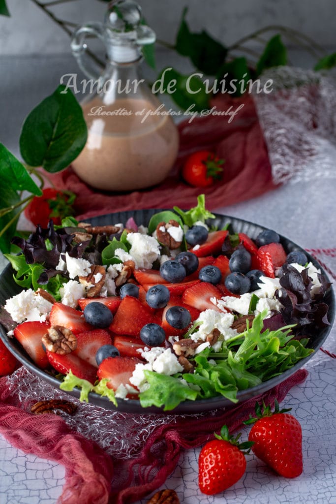 A colorful summer salad in a black bowl, featuring fresh green and purple lettuce, sliced strawberries, whole blueberries, crumbled feta cheese, and chopped pecans. In the background, a glass bottle of creamy strawberry vinaigrette sits on a red cloth, with whole strawberries and leafy green plants adding a fresh, decorative touch