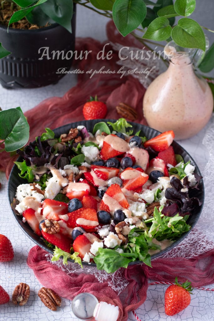 A vibrant summer salad served in a black bowl, featuring fresh mixed greens, sliced strawberries, blueberries, crumbled feta cheese, and chopped pecans, drizzled with a creamy strawberry vinaigrette. A glass bottle of vinaigrette and whole strawberries are placed nearby, with decorative greenery and fabric in the background