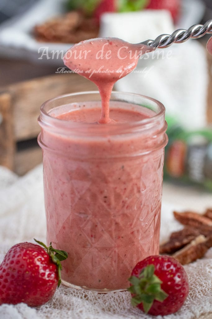A glass jar filled with creamy pink strawberry vinaigrette, placed on a textured white and red cloth with fresh strawberries scattered around. In the blurred background, a vibrant strawberry and feta salad is visible, adding a fresh and appetizing touch to the scene
