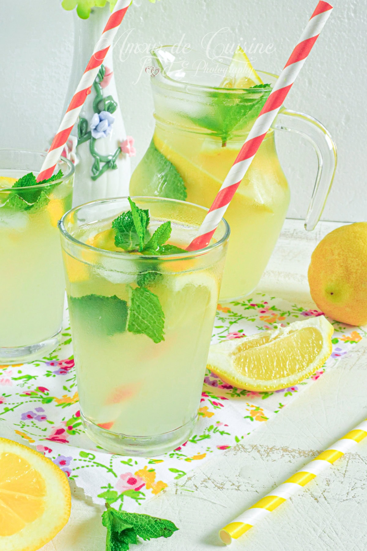 Verres de citronnade maison bien fraîche, garnis de rondelles de citron, de feuilles de menthe et de pailles rayées rouges et blanches, posés sur une nappe fleurie à côté d’une carafe de limonade, de citrons frais et de quartiers de citron, dans un décor lumineux et estival