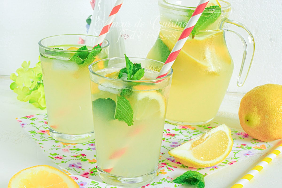 Verres de citronnade maison bien fraîche avec des rondelles de citron, des feuilles de menthe et des pailles rayées rouges, accompagnés d’une carafe de limonade, d’un citron entier, d’un quartier de citron et de quelques feuilles de menthe, le tout présenté sur une nappe fleurie dans un décor estival lumineux