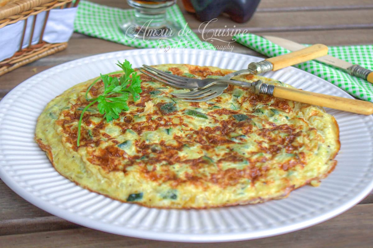 Omelette espagnole aux courgettes bien dorée, servie entière sur une assiette blanche avec une fourchette, un couteau rustique et une touche de persil frais – recette maison photographiée en extérieur.