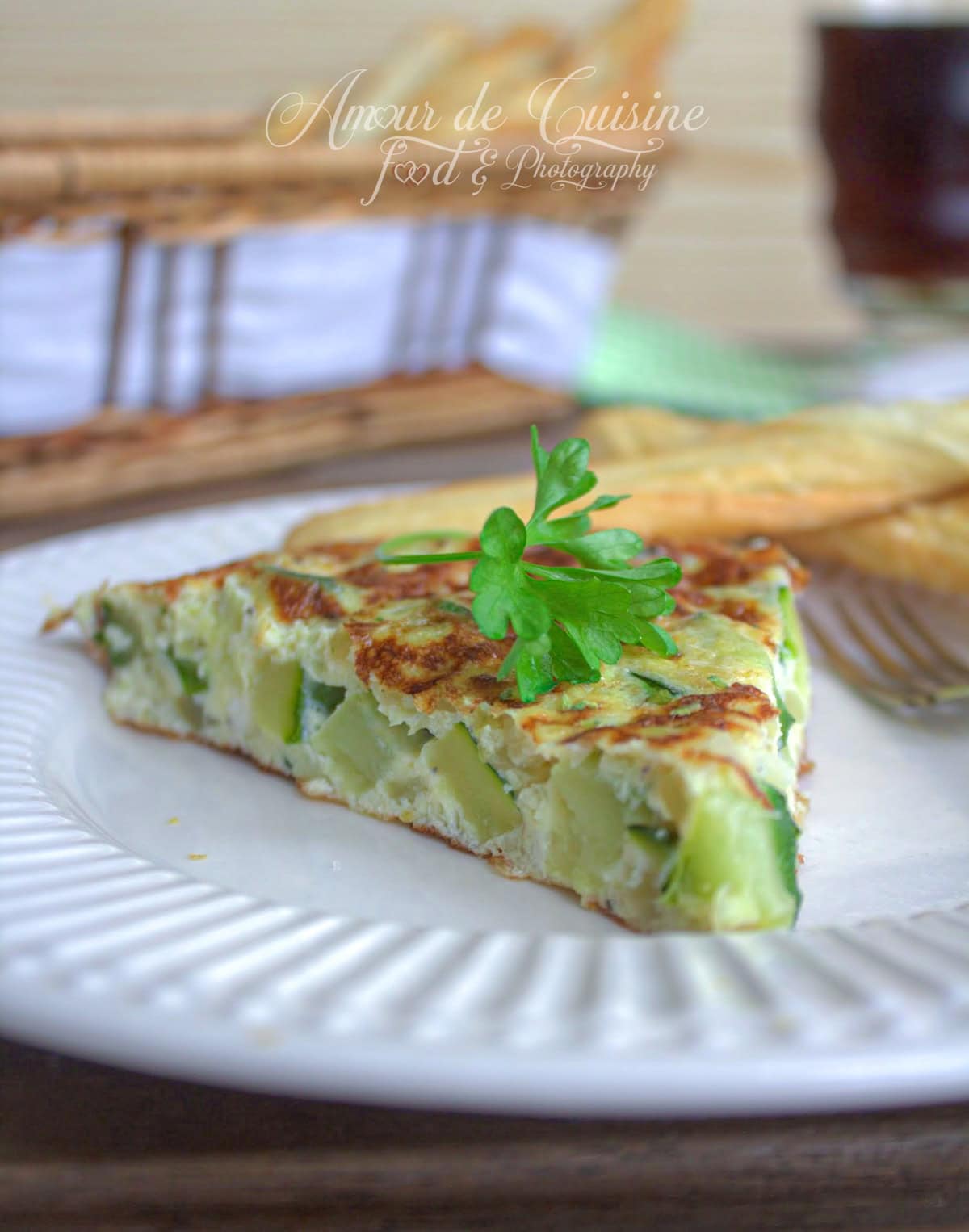 Tranche d’omelette espagnole aux courgettes servie sur une assiette blanche, décorée d’un brin de persil, accompagnée de pain et d’un verre en arrière-plan – recette maison photographiée par Amour de Cuisine