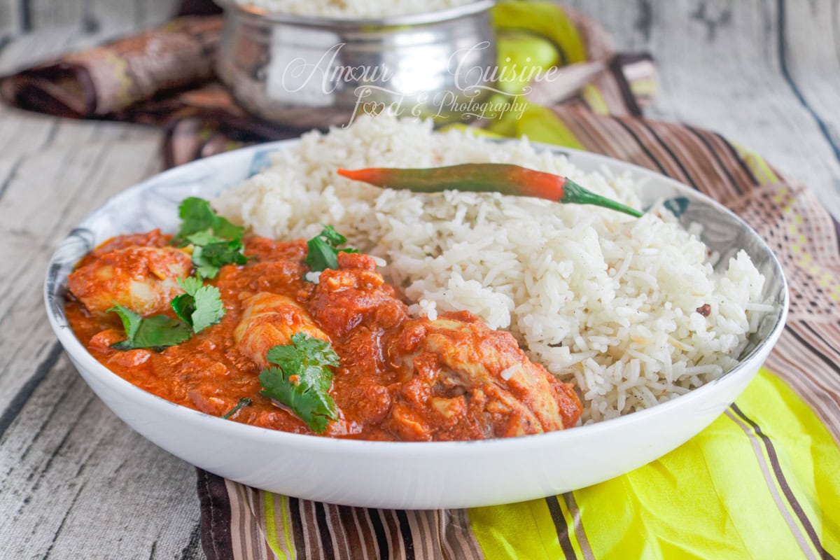 Plate of chicken tikka masala served with basmati rice, garnished with fresh coriander and a red chili pepper, spicy Indian dish presented on a rustic table.