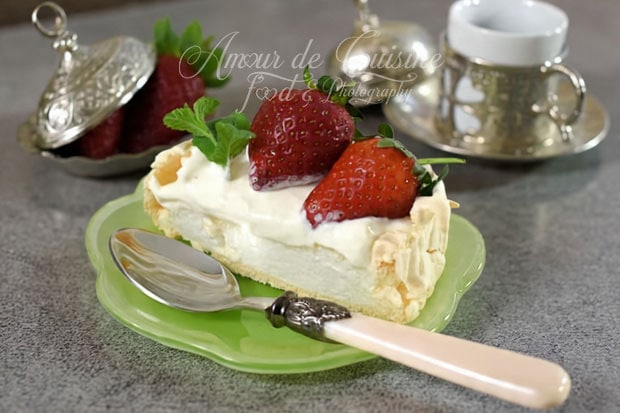 Slice of pavlova topped with whipped cream, fresh strawberries, and mint, served on a small green plate with a vintage spoon, with a cup and a dish of strawberries in the background.