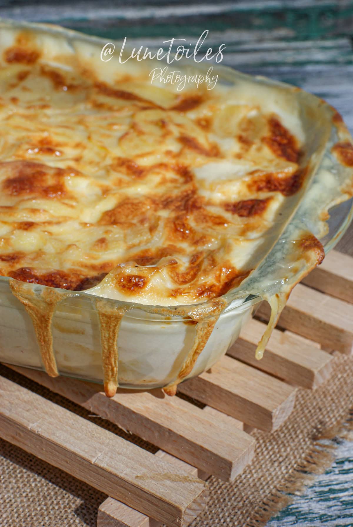 Gratin dauphinois maison, bien doré et crémeux, débordant légèrement d’un plat en verre, avec une croûte fondante et appétissante, posé sur un dessous-de-plat en bois rustique