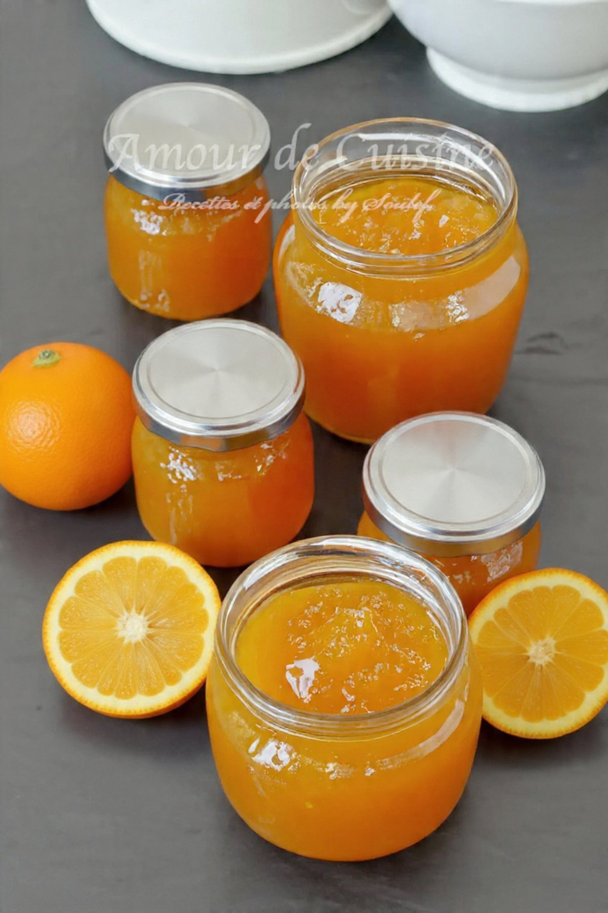 Jars of homemade orange jam on a grey countertop, with one whole orange and two orange halves placed around the jars.