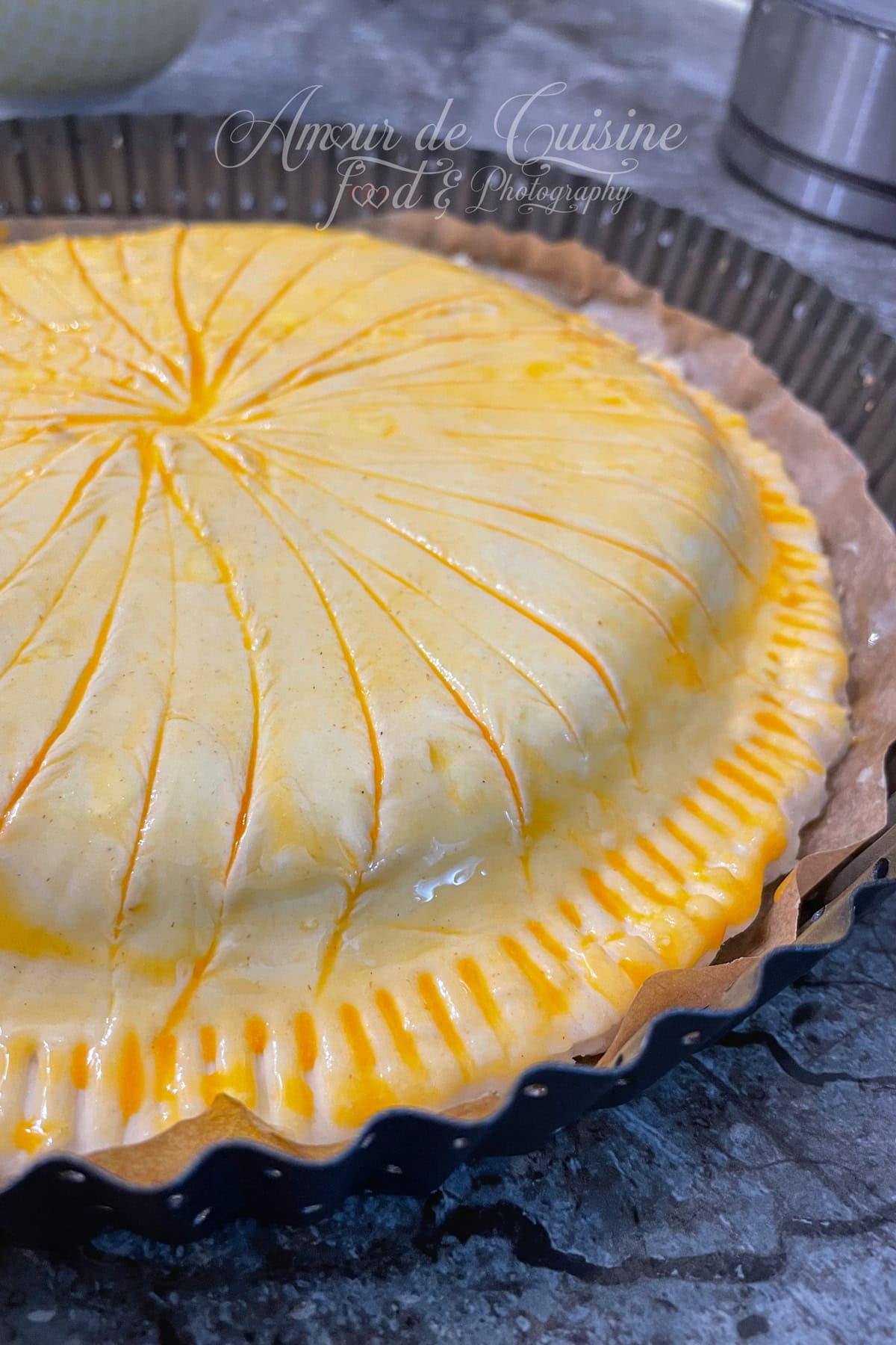 Unbaked Galette des Rois in puff pastry, brushed with egg yolk and scored with knife lines, crimped edges, set on parchment paper in a fluted pan before baking.