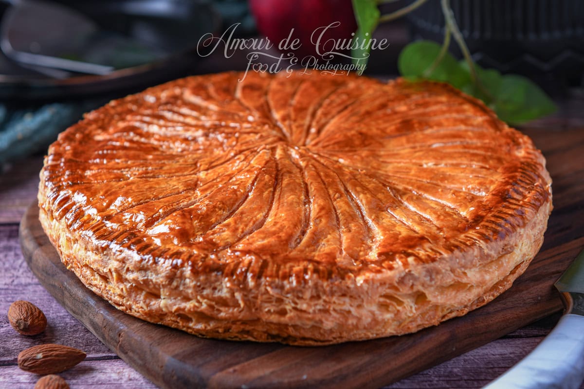 Whole Galette des Rois with deeply golden, glossy puff pastry, scored with a spiral knife pattern, set on a wooden board with a few almonds around it.