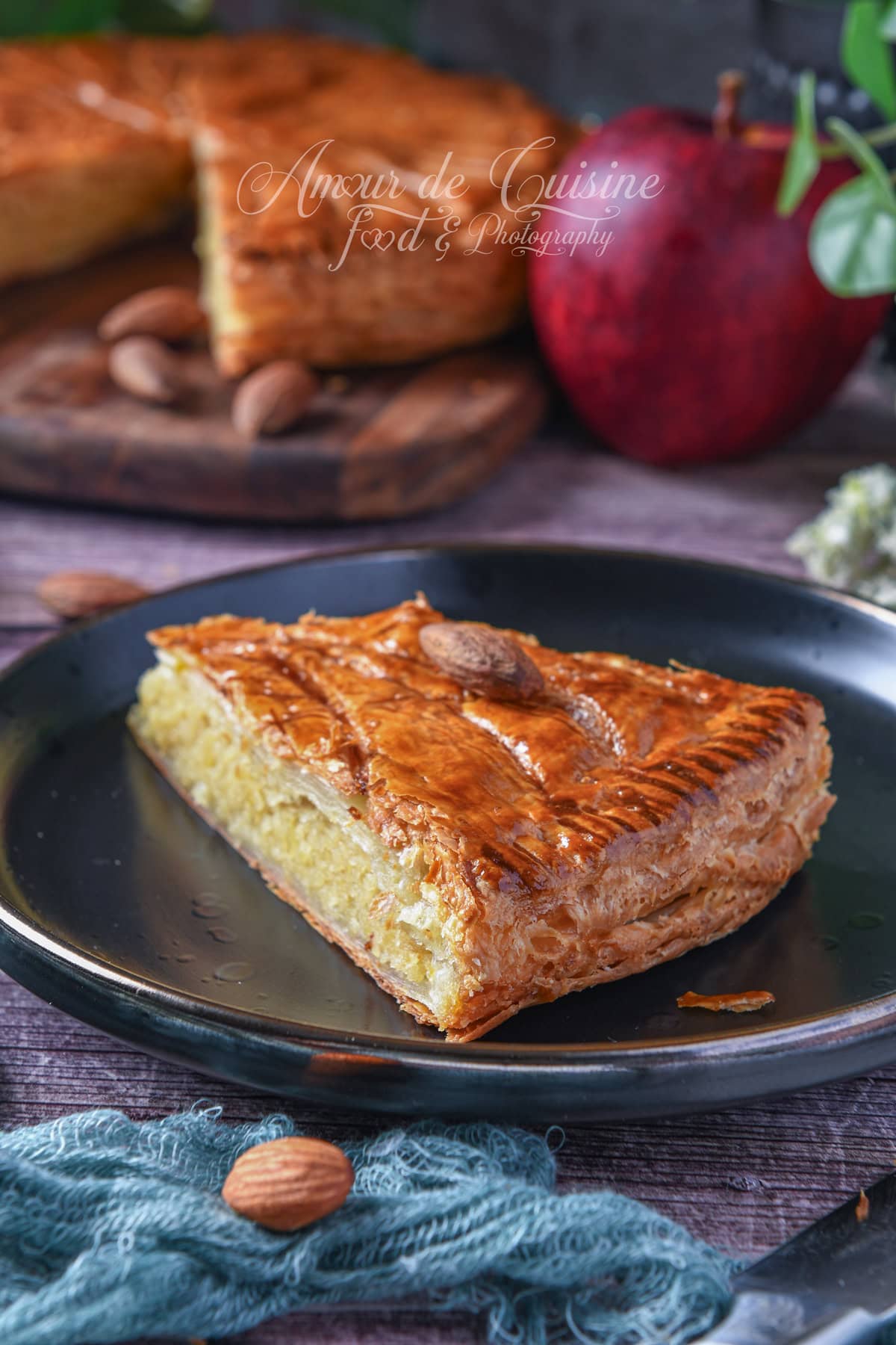 Slice of golden puff pastry Galette des Rois filled with almond cream, served on a black plate with an almond on top; the whole galette is blurred in the background and a red apple sits on the table.